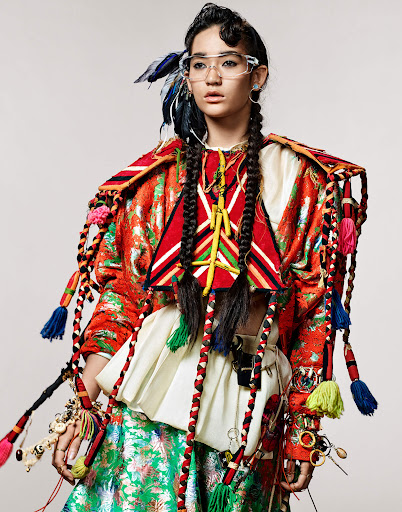 Portrait of Asian female fashion model wearing colorful ethnic-inspired clothing, eyeglasses, and braided hair against a neutral background.