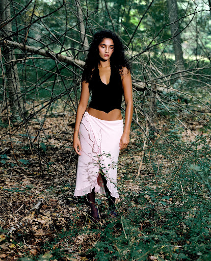 Woman with long curly hair, wearing a black top and light pink skirt, poses in an outdoor forest setting with trees and fallen branches.