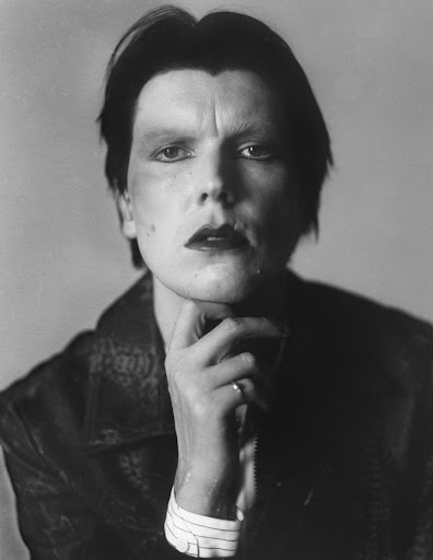Monochrome portrait of a male adult with dark hair and makeup, wearing patterned jacket, hand at chin, against a muted grey backdrop.