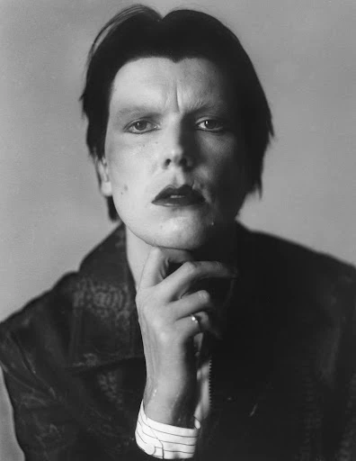 Monochrome portrait of a male adult with dark hair and makeup, wearing patterned jacket, hand at chin, against a muted grey backdrop.