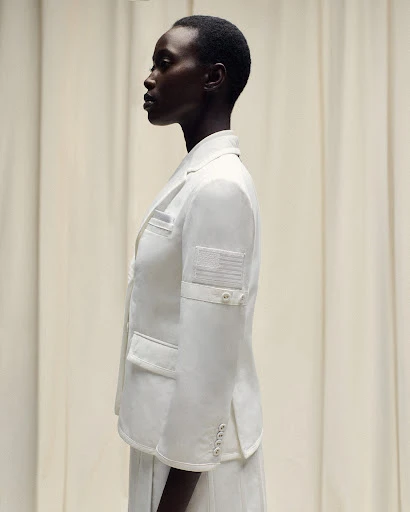 Profile of a dark-skinned woman in a white blazer with an American flag patch, set against a neutral-toned curtain backdrop.