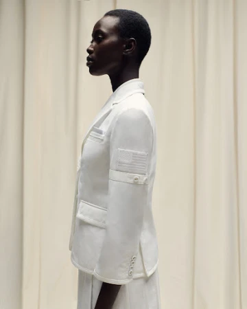 Profile of a dark-skinned woman in a white blazer with an American flag patch, set against a neutral-toned curtain backdrop.