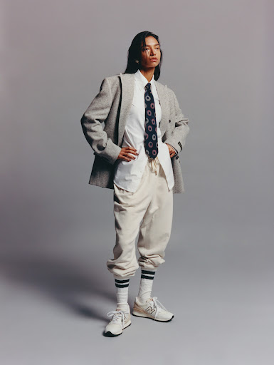 Portrait of a young woman posing in a grey herringbone blazer, white collared shirt, navy tie, beige sweatpants, socks, and sneakers against a grey background.