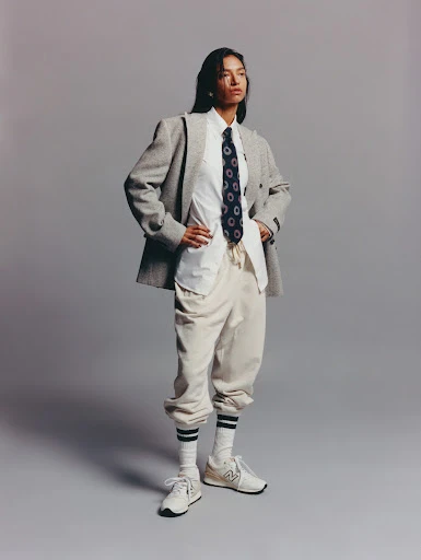 Portrait of a young woman posing in a grey herringbone blazer, white collared shirt, navy tie, beige sweatpants, socks, and sneakers against a grey background.
