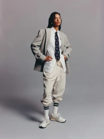 Portrait of a young woman posing in a grey herringbone blazer, white collared shirt, navy tie, beige sweatpants, socks, and sneakers against a grey background.