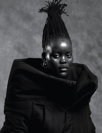 Monochrome portrait of a Black woman with tall sculpted dreadlocks wearing a heavy black hooded garment against a textured grey studio backdrop