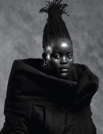 Monochrome portrait of a Black woman with tall sculpted dreadlocks wearing a heavy black hooded garment against a textured grey studio backdrop