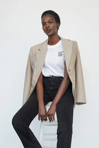 Studio shot of a Black female model wearing a blazer, tee shirt, and jeans, sitting on a white stool against a white backdrop.