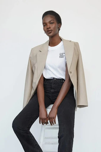 Studio shot of a Black female model wearing a blazer, tee shirt, and jeans, sitting on a white stool against a white backdrop.