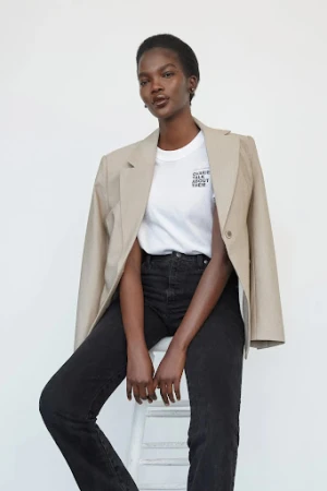 Studio shot of a Black female model wearing a blazer, tee shirt, and jeans, sitting on a white stool against a white backdrop.