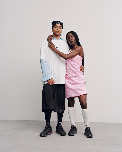 Two Black adults in studio setting showcasing sneakers and sportswear. The woman wears a pink dress while the man wears a white t-shirt.