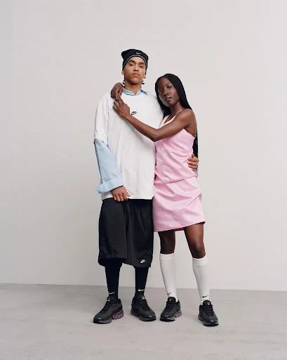 Two Black adults in studio setting showcasing sneakers and sportswear. The woman wears a pink dress while the man wears a white t-shirt.