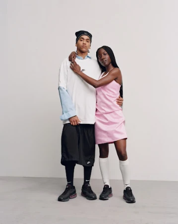 Two Black adults in studio setting showcasing sneakers and sportswear. The woman wears a pink dress while the man wears a white t-shirt.