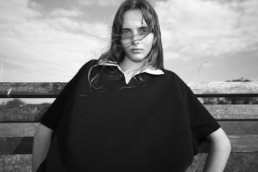 Black and white portrait of a young woman with hair blowing across her face, seated outdoors on a wooden park bench.