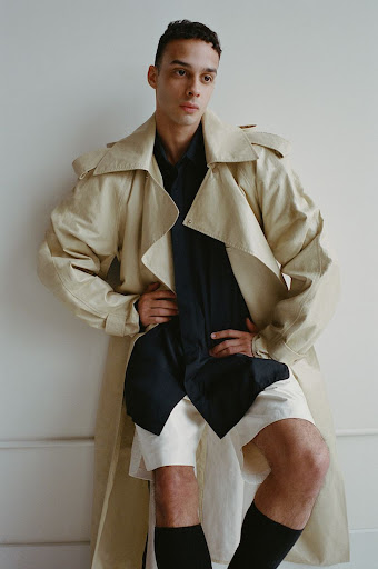 A fashionable young adult stands indoors, wearing a tan trench coat over a black shirt and white shorts, plus black socks, against a white wall.