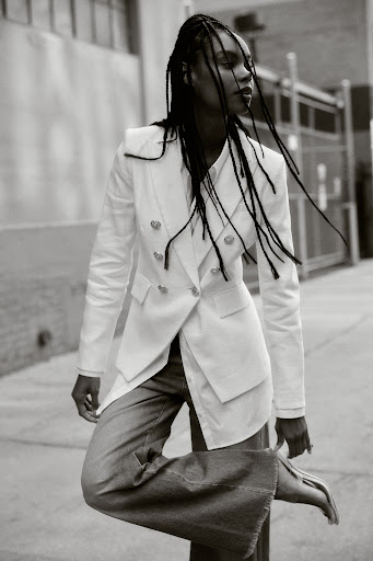 Black and white editorial portrait of a woman with braided hair wearing a white blazer and wide-leg jeans outdoors.