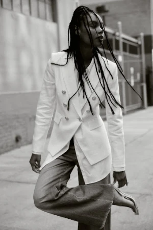 Black and white editorial portrait of a woman with braided hair wearing a white blazer and wide-leg jeans outdoors.