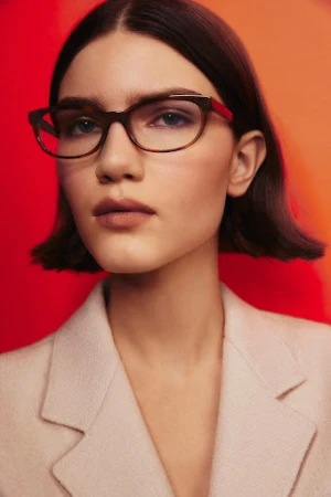 Close-up portrait of a woman wearing tortoiseshell glasses and a light blazer against a bold red and orange background.