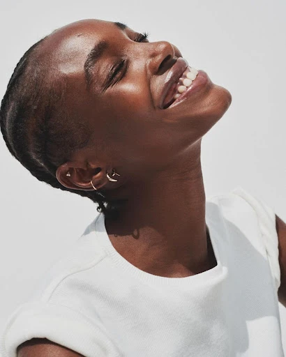 Portrait of a Black woman with short hair and bright white teeth, smiling and looking up with a light background. Jewelry visible.