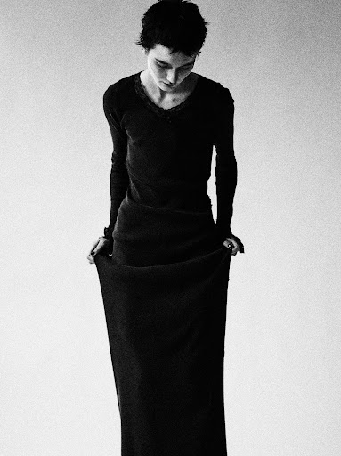 A person in a black long-sleeve shirt and skirt stands in a white studio, holding the skirt hems while looking down in a medium shot.
