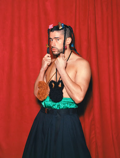 Bad Bunny poses against a red curtain, wearing a green top, black skirt, and unique brown and black fuzzy crocheted shoulder bags.