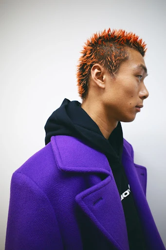 Profile shot of a stylish Asian male wearing a purple overcoat and black hoodie, showcasing an orange spiky hairstyle against a white backdrop.