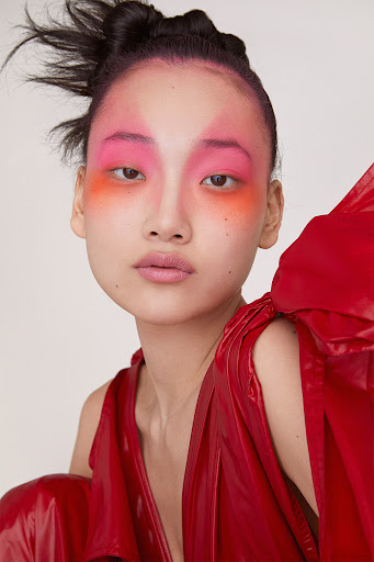 Close-up of a fashion model with bright pink and orange makeup on her face and upper body in an indoor photo studio setting.