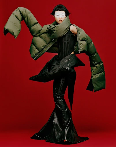 Asian woman in avant-garde fashion poses against a red background, wearing an olive green puffer jacket and a dramatic black vinyl dress.