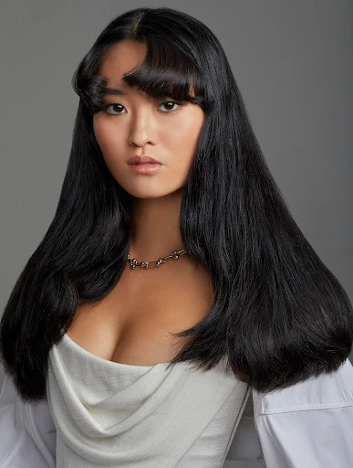 Portrait of a young Asian woman with long, dark hair wearing a white top and silver necklace, captured in a photo studio setting.