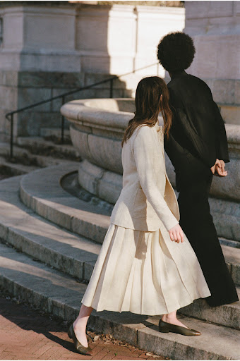 Two models walk on stone steps. One is an African American woman wearing all black. The other is a Caucasian woman wearing a skirt suit.