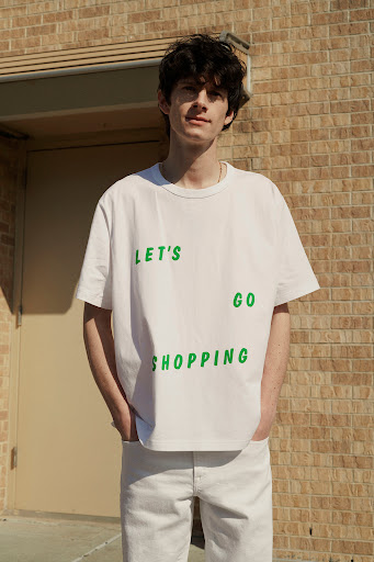 A young adult male posing in front of a brick wall, wearing a white tee with 'LET'S GO SHOPPING' printed on it, and white jeans.