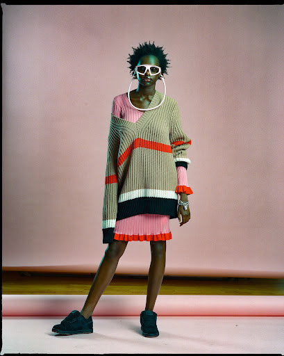 A fashionable Black woman stands in a photo studio wearing a beige knit sweater dress, black shoes, white sunglasses and hoop earrings.
