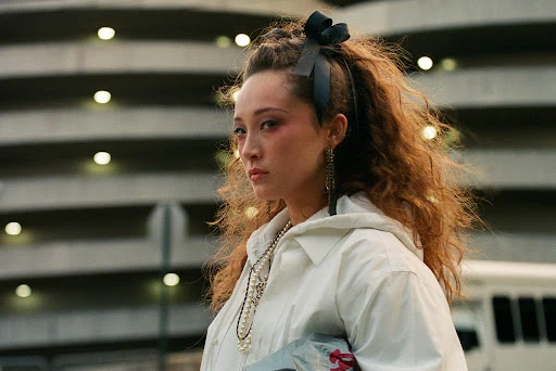 Portrait of a woman with curly hair and a white jacket, accented with eye-catching pink eye makeup, posing outdoors against a parking garage.