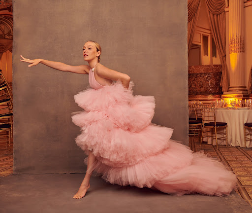 Woman in a pink tiered tulle gown poses barefoot in a photo studio with a neutral background and a vintage-style hotel ballroom set piece.