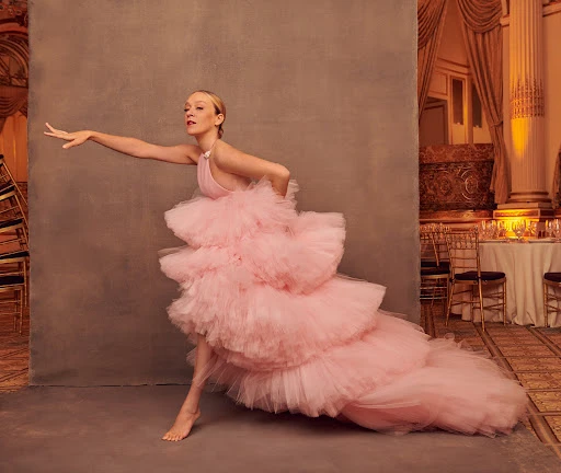 Woman in a pink tiered tulle gown poses barefoot in a photo studio with a neutral background and a vintage-style hotel ballroom set piece.
