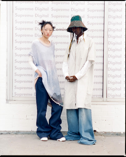 Two fashion models pose outdoors against a wall with 'Supreme Digital' text. The model on the left wears a light blue poncho and blue jeans.