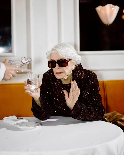 Elderly woman in sunglasses sits at restaurant table as waiter pours water from a bottle into a glass, creating a lifestyle portrait.