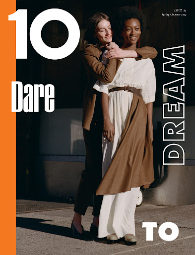Two women posing together outdoors in sunlight, one wearing a brown blazer and trousers and the other a white dress with a brown apron skirt.
