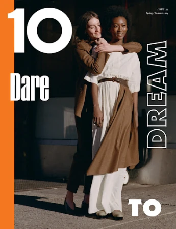 Two women posing together outdoors in sunlight, one wearing a brown blazer and trousers and the other a white dress with a brown apron skirt.