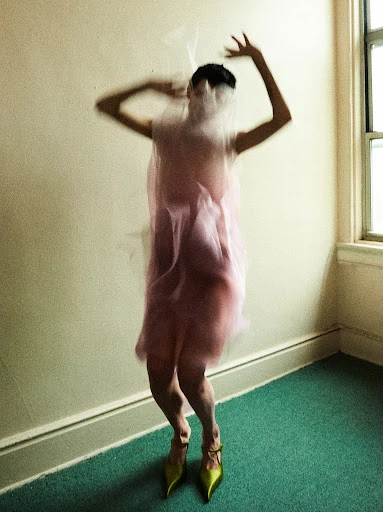 A blurred woman in pink dress and yellow heels stands in a room, mostly obscured by a transparent veil or fabric.