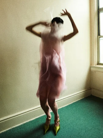 A blurred woman in pink dress and yellow heels stands in a room, mostly obscured by a transparent veil or fabric.
