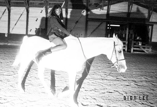 Black and white shot of a woman riding a white horse in an indoor arena, wearing a jacket, shorts, and boots with arena structures in the background.