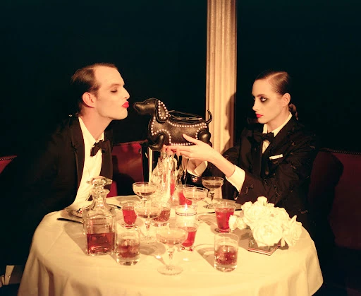 Two models in tuxedos at a table laden with drinks and a dachshund purse, set against a dark background and dramatic lighting.