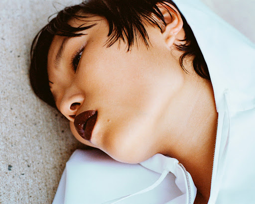 Close-up portrait of a young woman with short dark hair and dark lipstick, lying on a gray surface wearing a white hooded jacket.