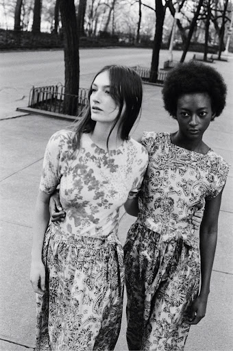 Black and white fashion shot of two women in matching paisley-print outfits with arms around each other on a city street.