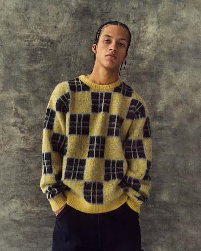 A young adult man in a yellow and black checkered sweater poses against a mottled grey backdrop, hands tucked into his pants.