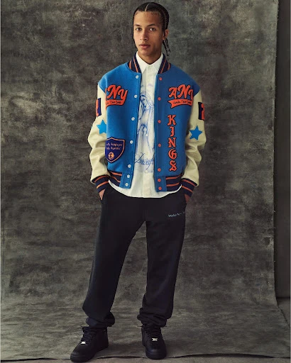 Male fashion model poses indoors wearing a letterman jacket, collared shirt, sweatpants, and sneakers in a studio setting.