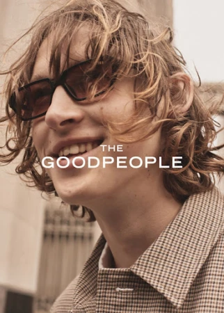 Close-up of a smiling adult male with blond hair and sunglasses, wearing a brown plaid jacket against an urban background.