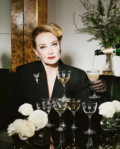 Elegant woman in a black suit pours champagne into a stemmed glass, surrounded by white flowers and a cabinet, in a luxurious interior.
