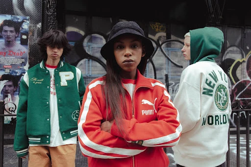 Three models in streetwear including a central woman in a red zip-up jacket against a graffitied urban street background.